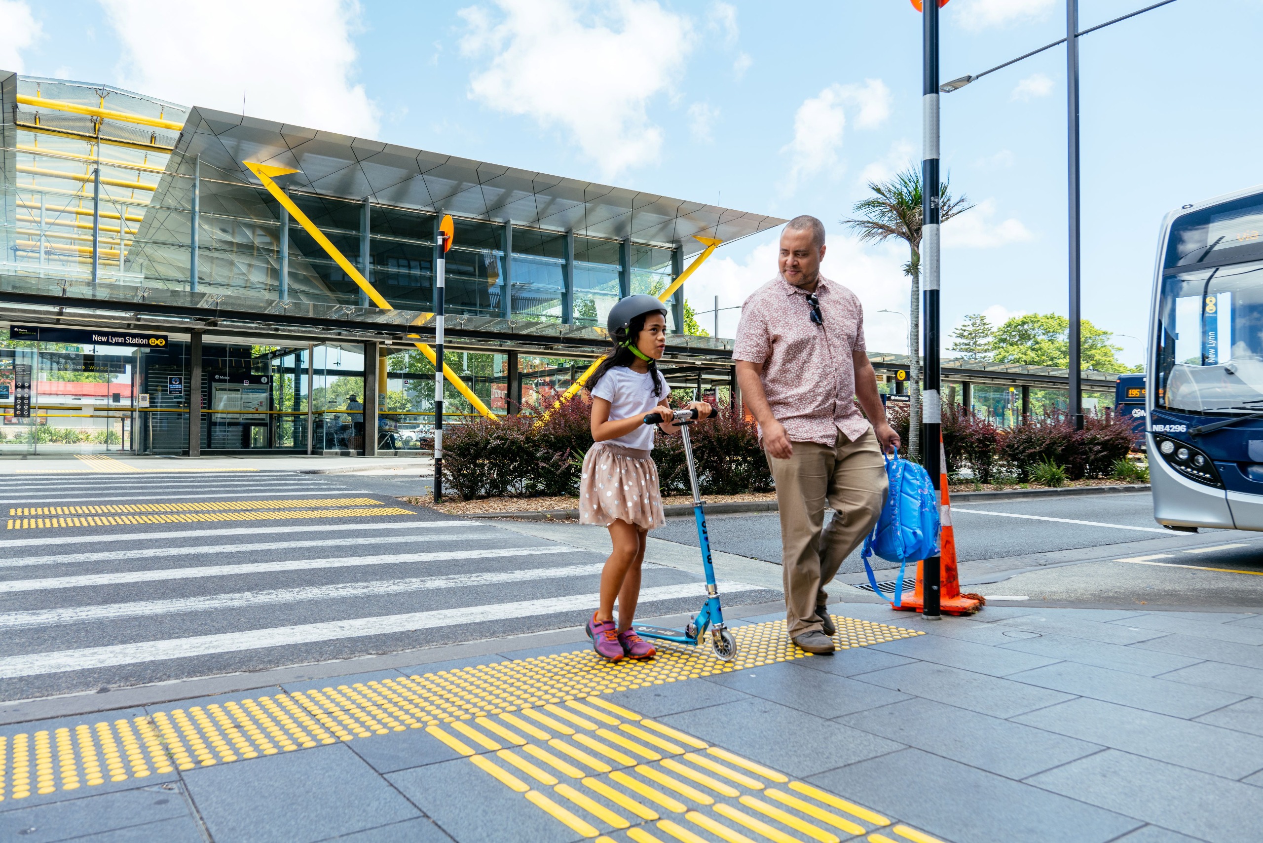 A parent carrying a backpack and a child pushing their scooter while crossing a pedestrian crossing